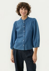 Jeansblus med rund krage, knäppning fram och volangärmar. Blått tyg med en mjuk textur. Litet kortare längd.