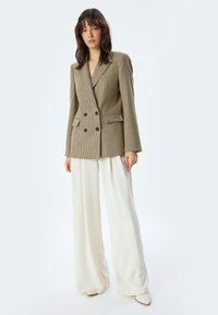 Blazer doppiopetto marrone a quadri con bottoni neri, abbinato a pantaloni bianchi a gamba larga e fluidi. Texture liscia e vestibilità strutturata.