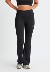 Mujer con leggings deportivos negros de talle alto y un sujetador deportivo negro, de pie con las manos en las caderas y zapatillas de correr plateadas.