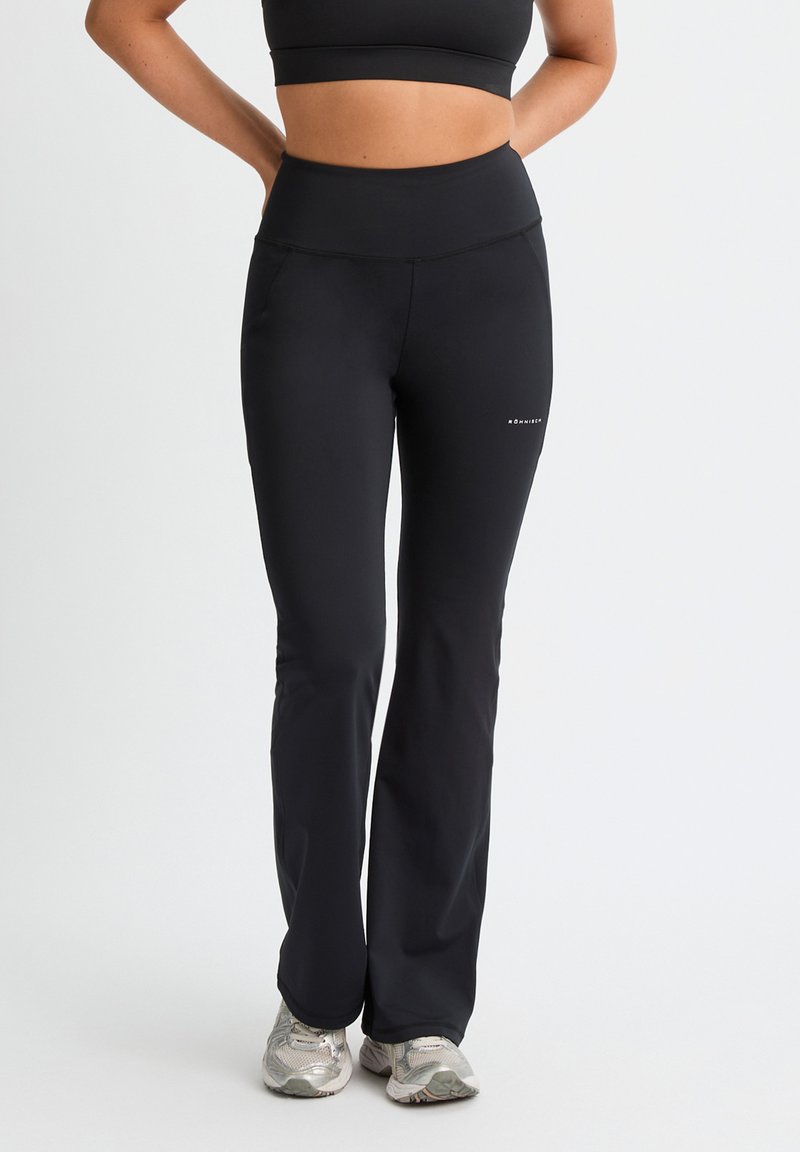 Mujer con leggings deportivos negros de talle alto y un sujetador deportivo negro, de pie con las manos en las caderas y zapatillas de correr plateadas.