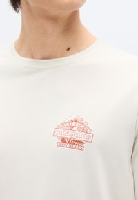 Κοντινό πλάνο προσώπου που φοράει εκρού μπλουζάκι με κόκκινο σχέδιο και τη φράση "Great Pacific Patagonia Iron Works" που απεικονίζει κυματιστό ωκεανό και βουνό.