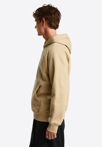 Seitenansicht eines Mannes, der einen beigefarbenen Kapuzenpullover mit Kängurutasche und schwarze Hosen trägt, vor einem schlichten hellen Hintergrund steht.