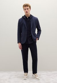 Blazer navy con cappuccio, camicia nera con bottoni, pantaloni navy e sneakers beige. Tessuto strutturato con tasche e coulisse sul cappuccio.