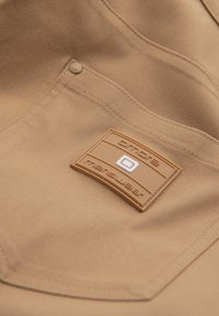 Gros plan d'une poche en tissu beige avec des bords cousus et un patch en caoutchouc rectangulaire intitulé "Dyne Rescuewear" avec un carré blanc central.