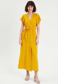 Yellow wrap dress made of lightweight fabric, featuring short ruffled sleeves, a tie belt, and a front slit. Worn with strappy heels.