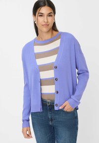 Lavenderfarbener, gerippter Cardigan mit lockerer Passform, ausgestattet mit braunen Knöpfen, kombiniert mit einem gestreiften Oberteil in Beige und Blau, getragen mit dunklen Jeans.