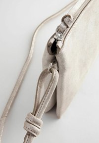 Beżowa skórzana torebka kopertówka z zamkiem błyskawicznym, regulowanym paskiem i dekoracyjnym węzłem na pasku. Gładka tekstura i minimalny design.