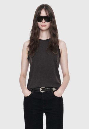 Woman with long brown hair wearing black sleeveless top, black pants, black belt, and large black sunglasses with hands in pockets.