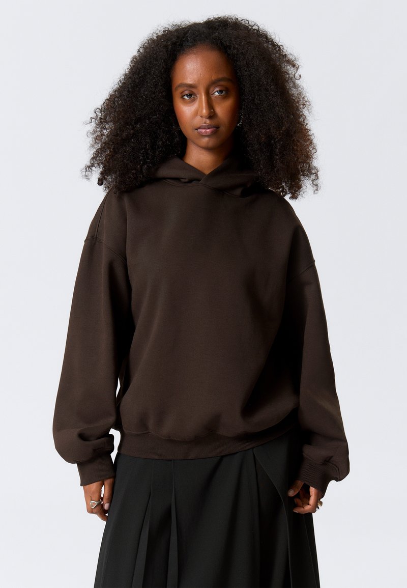 Sudadera con capucha marrón hecha de un tejido suave, con hombros caídos y puños acanalados, combinada con una falda negra plisada.