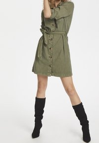 Olivově zelené šaty s knoflíky a stahovací šňůrkou, které mají dvě boční kapsy a ohrnuté rukávy. Nosené s černými volnými vysokými kozačkami.