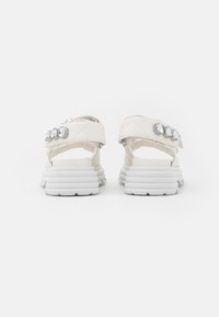 Sandales blanches avec semelles en caoutchouc multicouches, présentant des brides synthétiques matelassées ornées de trois pierres précieuses transparentes de chaque côté.