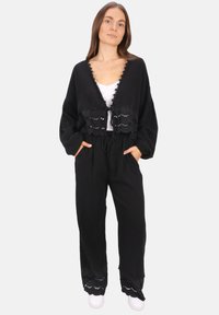 Schwarze, kurz geschnittene Strickjacke mit gewellten Kanten, kombiniert mit passenden lockeren Hosen mit Spitzenakzenten. Ein weißes Oberteil und Turnschuhe runden das Outfit ab.