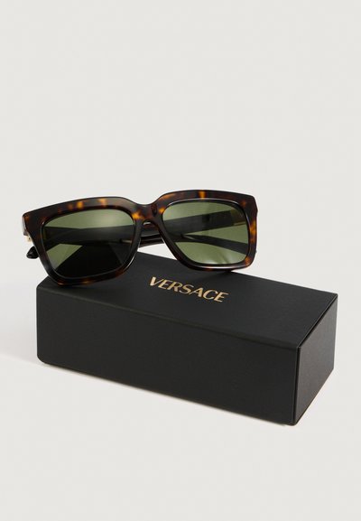 Occhiali da sole rettangolari in tartaruga con lenti verde scuro appoggiati su un astuccio nero della Versace.