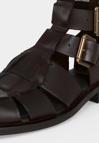 Σανδάλι από σκούρο καφέ δέρμα με πλεγμένους ιμάντες και διπλές αγκράφες σε χρυσό τόνο σε μαύρη σόλα.