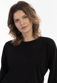 Fekete, hosszú ujjú felső, laza szabással és kerek nyakkivágással, sima textúrával. A háttér sima fehér, hangsúlyozva a ruhadarabot.