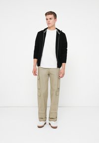 Nuori mies seisoo mustassa vetoketjullisessa hupparissa, valkoisessa t-paidassa, beigekalvoisissa cargo-housuissa ja valkoisissa lenkkareissa tasaisessa valkoisessa taustassa.