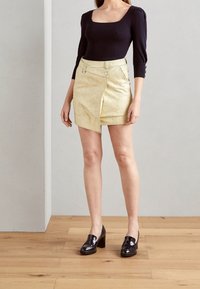 Schwarzes, geripptes Langarmoberteil kombiniert mit einem metallisch goldenen Minirock mit asymmetrischem Saum und Vorderreißverschluss. Schwarze Lack-Loafer.