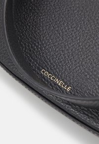 Gros plan d’un sac en cuir noir texturé montrant le logo doré Coccinelle imprimé à l’intérieur du bord arrondi.