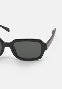 Ochelari de soare negri cu lentile dreptunghiulare, rame groase din plastic și margini moi. Lentilele de culoare gri închis oferă protecție UV. Brațele subțiri negre se extind spre afară.