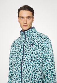 Jachetă din fâș cu un print de leopard aqua și bleumarin, fermoar complet, guler înalt și un mic logo Nike bleumarin pe pieptul stâng.