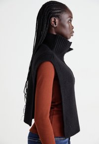 Profil einer Frau mit geflochtenen Haaren, die einen schwarzen ärmellosen Strickumhang über einem rostfarbenen Langarmoberteil und blauen Jeans trägt, vor einem weißen Hintergrund.