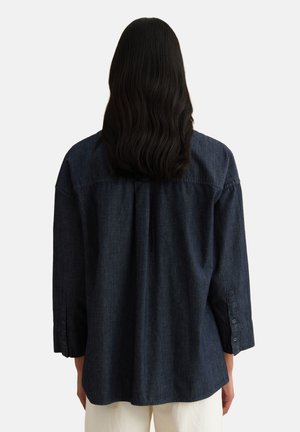 Woman with long black hair wearing a loose dark denim long-sleeve shirt and light-colored pants, shown from the back.