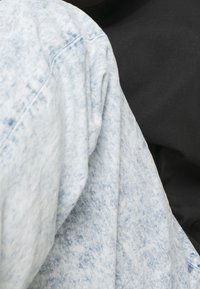 Chaqueta de mezclilla en azul claro con una textura desgastada, que presenta un corte ligeramente oversized, junto a una prenda negra en el fondo.