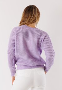 Donna con capelli lunghi che indossa un maglione di lana lilla e pantaloni bianchi, vista di spalle su uno sfondo neutro.