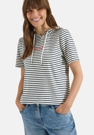 Kvinde smilende iført en kortærmet hvid hættetrøje med sorte vandrette striber og blå jeans, med hænderne i lommerne mod en ensfarvet baggrund.