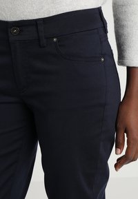 Námořnické modré kalhoty vyrobené z bavlněné směsi, které mají přední kapsu, jemné prošívání a hladkou texturu. Zapínání na kovový knoflík.