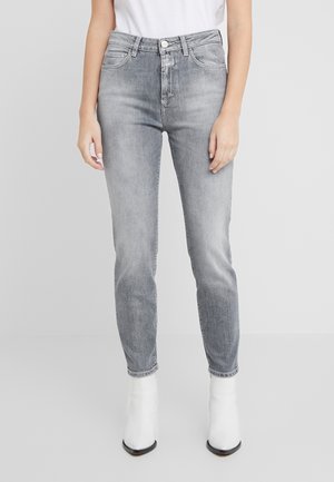 Personne portant un jean gris délavé taille haute et des bottines blanches, debout les bras relâchés le long du corps devant un fond uni.