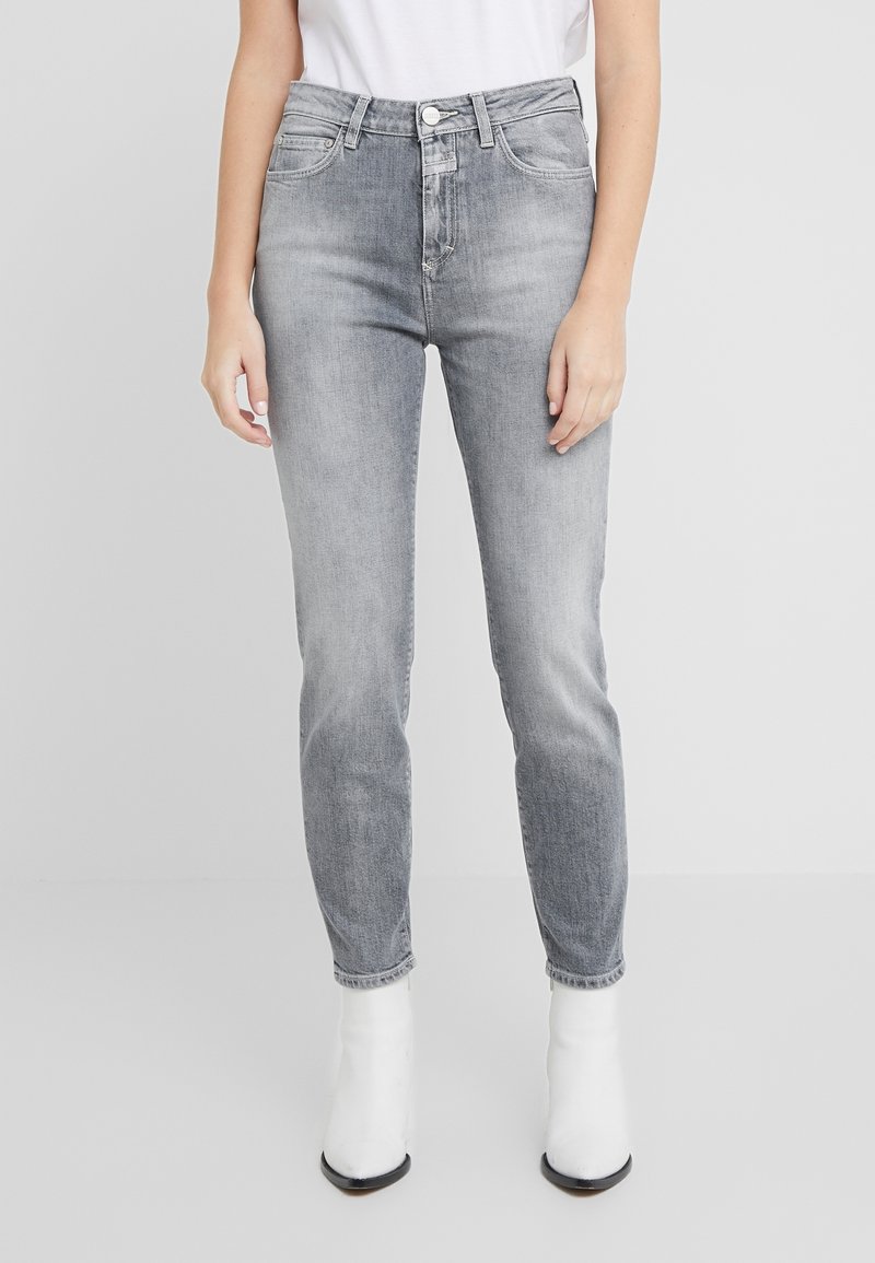 Personne portant un jean gris délavé taille haute et des bottines blanches, debout les bras relâchés le long du corps devant un fond uni.