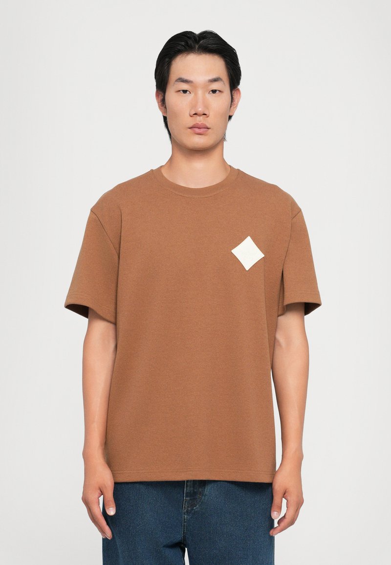 Brown short-sleeve t-shirt with a round neckline and a diamond-shaped logo patch in cream. Fabric appears soft and slightly textured.