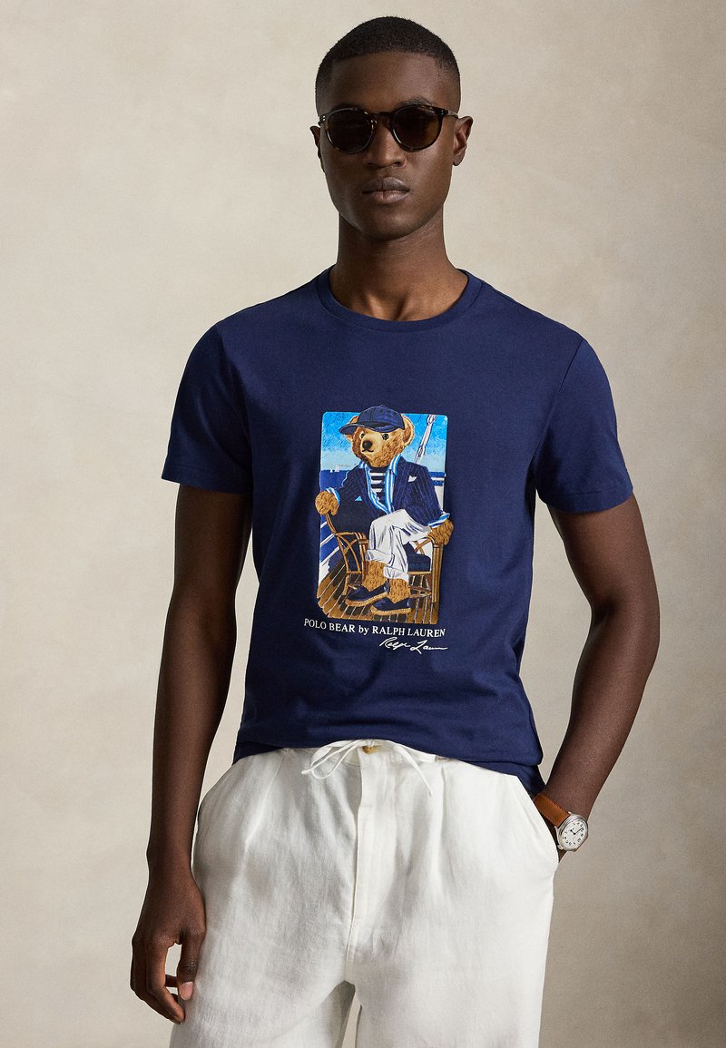Homme portant des lunettes de soleil foncées, un t-shirt bleu marine avec un ours en blazer imprimé et un pantalon blanc à cordon devant un fond beige uni.