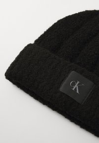 Musta neulepipo, jossa on teksturoitu pinta, taitettu reunus ja musta nahkalaatta, jossa on kohokuvioitu logo. Pehmeä ja lämmin materiaali.