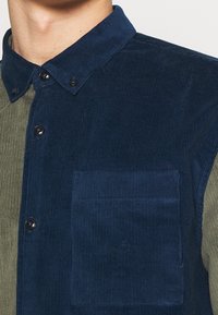 Pellavapaita, jossa on vaihteleva väripaletti tummansinistä ja oliivinvihreää. Huomiota herättävät yksityiskohdat ovat napillinen edusta ja vasemman rinnan tasku.