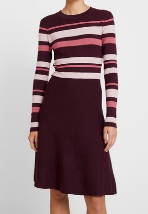 Kobieta ubrana w sukienkę do kolan z jednolitą, ciemnofioletową spódnicą oraz długim rękawem topu w różowe i fioletowe poziome paski.
