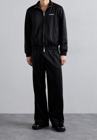 Black zip-up tracksuit with ribbed cuffs and waistband, featuring a logo on the chest. Paired with glossy black shoes on a textured background.
