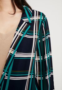 Námořnický kostkovaný blejzr s tyrkysovými, bílými a žlutými pruhy. Lehčí látka s klopovým límcem a otevřeným předním dílem.