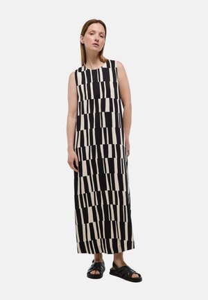 Femme portant une robe longue sans manches à motif géométrique noir et blanc avec des sandales noires ouvertes aux orteils sur un fond uni.