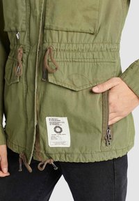 Veste de style militaire verte avec poches, cordons de serrage et tissu texturé. Comprend une étiquette sur l'ourlet et une poche zippée.