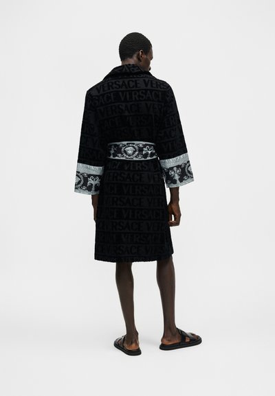 Άνδρας στέκεται, φορώντας μαύρη ρόμπα Versace με ζώνη και μανσέτες με σχέδιο, συνδυασμένη με μαύρα σανδάλια slide, σε απλό φωτεινό φόντο.