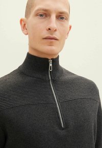 Dunkelgrauer Reißverschluss-Pullover mit gerippter Textur und horizontalen Streifen am oberen Ende. Verfügt über einen hohen Kragen und silberne Reißverschlussbeschläge.