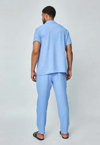 Chemise bleu clair, à manches courtes et col, avec un pantalon léger assorti. Les deux sont en tissu texturé. Porté avec des sandales noires.