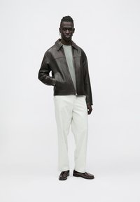 Brun skinnjakke med glidelås, grå skjorte under, kombinert med lyse bukser og mørke sko. Enkelt, avslappet passform.