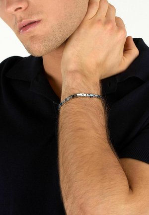 Uomo con camicia nera tiene il polso mostrando un braccialetto in metallo argento e blu al braccio sinistro.