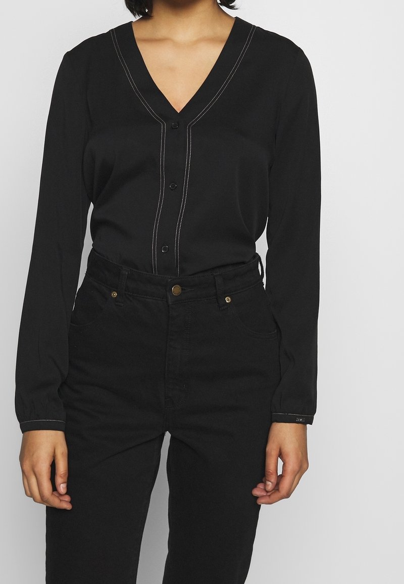 Camicia nera con bottoni, scollo a V, maniche lunghe e cuciture a contrasto; abbinata a pantaloni neri a vita alta con tasche standard.