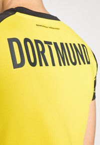 Maillot de sport jaune arborant « DORTMUND » en lettres noires en gras au dos. Le matériau est texturé avec des accents noirs sur les épaules.