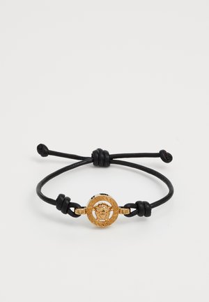 Bracelete de couro com um medalhão redondo dourado, apresentando um design detalhado de um rosto, segurado com nós de corda preta ajustáveis.
