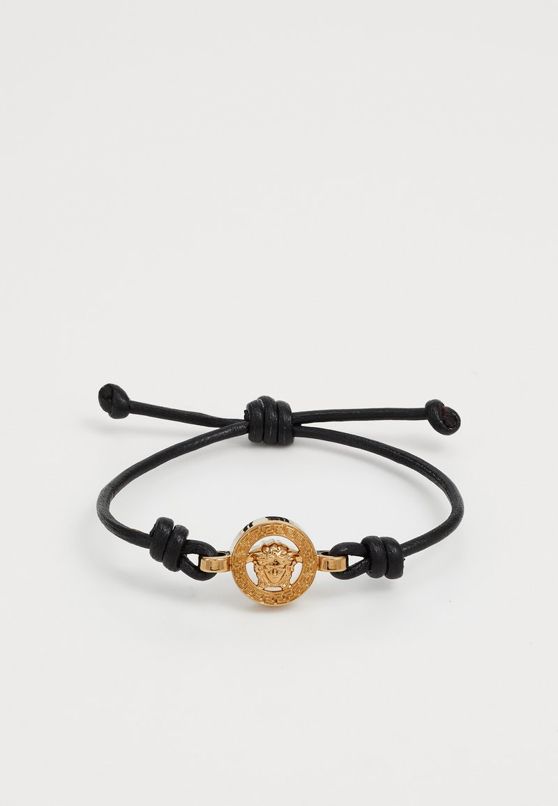 Bracciale in pelle con un medaglione rotondo d'oro caratterizzato da un design dettagliato di un volto, fissato con nodi regolabili in corda nera.